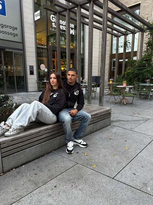 Two models wearing black oversized Clover at Night hoodies by R3CEIVED sitting outdoors on a bench in an urban setting