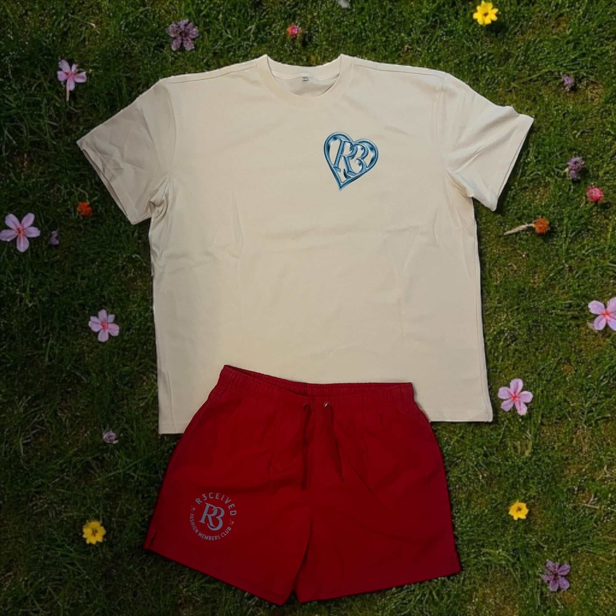 Men's red R3 Fashion Members Club swimming trunks with pockets and elastic waistband, paired with a cream t-shirt, displayed on grass with flowers.