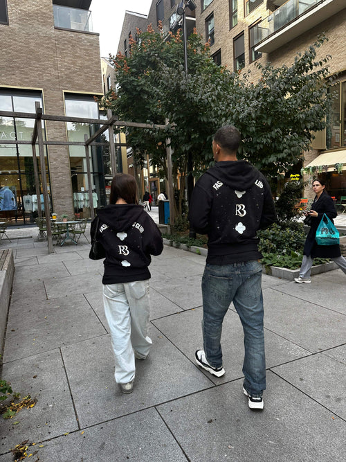 Two people wearing black Clover at Night hoodies with R3 logo and clover designs on the back walking outdoors in an urban setting
